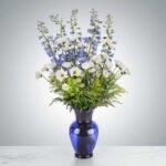 A tall floral arrangement in a deep blue glass vase featuring blue delphiniums, white daisies, and green foliage against a grey gradient background.