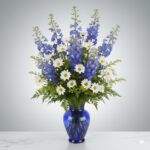 A tall, elegant floral arrangement featuring vibrant blue delphiniums, white daisies with yellow centers, and lush green ferns in a translucent cobalt blue glass vase, set against a neutral gray background.
