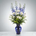 A vertical floral arrangement featuring tall blue delphiniums, white daisy poms, and green filler plants in a translucent cobalt blue glass vase.