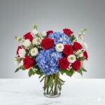 A lush floral arrangement in a glass vase featuring red roses, blue hydrangeas, and white lisianthus on a marble surface.