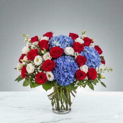 A lush and patriotic floral arrangement in a glass vase featuring vibrant red roses, large blue hydrangeas, and delicate white lisianthus or spray roses, set against a neutral grey background on a marble surface.