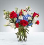 A patriotic floral arrangement in a clear glass vase featuring red roses, blue hydrangeas, and white lisianthus with green foliage on a marble surface.