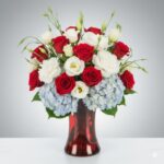 A lush floral arrangement in a red glass vase featuring red roses, white lisianthus, and light blue hydrangeas against a neutral grey gradient background.