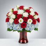 A patriotic "Americana" floral arrangement featuring red roses, white lisianthus, and light blue hydrangeas in a translucent red glass vase, set against a neutral gray gradient background.