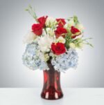 A patriotic-themed flower bouquet featuring red roses, white lisianthus, and light blue hydrangeas arranged in a translucent ruby-red glass vase.