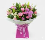 A lush and vibrant floral bouquet featuring a mix of pink roses, purple daisies, pink carnations, and white chrysanthemums with green lily buds, elegantly wrapped in white paper and presented in a bright pink gift box with a checkered pink ribbon.