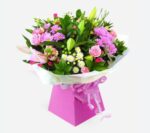 A lush, vibrant flower bouquet featuring pink roses, purple chrysanthemums, and white daisies accented with green foliage, presented in a pink decorative box with a checkered ribbon.