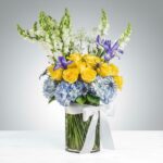 A vibrant floral arrangement in a glass vase featuring yellow roses, blue hydrangeas, purple irises, and white snapdragons, decorated with several black plastic spiders for a Halloween theme.