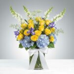 A vibrant floral arrangement in a glass vase featuring yellow roses, blue hydrangeas, purple irises, and white snapdragons, decorated with small black plastic spiders.