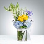 A vibrant floral arrangement in a glass cylinder vase featuring white snapdragons, yellow roses, blue hydrangeas, and purple iris, finished with a white satin bow.