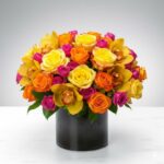 A lush, vibrant flower arrangement featuring a dense mix of yellow roses, orange roses, hot pink spray roses, and yellow cymbidium orchids in a sleek black cylindrical glass vase.