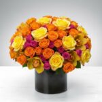 A lush and vibrant floral arrangement featuring a dense cluster of yellow, orange, and hot pink roses, mixed with yellow orchids, presented in a sleek cylindrical black glass vase on a white marble surface.