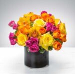 A vibrant and dense floral arrangement featuring yellow roses, orange roses, hot pink spray roses, and yellow cymbidium orchids in a sleek black cylindrical vase on a white marble surface.