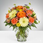 A vibrant and colorful "Carnival" mixed flower arrangement in a clear glass ginger vase, featuring orange roses, pale yellow gerbera daisies, orange carnations, pink alstroemeria, and purple aster accents with lush green ferns.