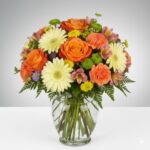 A vibrant and colorful floral bouquet in a clear glass vase, featuring orange roses, pale yellow gerbera daisies, orange carnations, pink alstroemeria, and green button poms, arranged with lush green ferns.