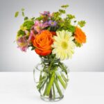 A vibrant carnival-themed flower arrangement in a clear glass vase featuring orange roses, a large pale yellow gerbera daisy, pink alstroemeria, and purple asters.
