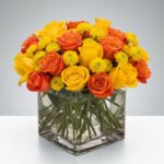 A vibrant bouquet of yellow and orange roses mixed with yellow button poms in a square glass vase filled with clear water.