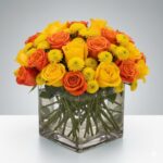 A vibrant floral arrangement featuring orange and yellow roses mixed with small yellow pompon chrysanthemums in a square clear glass vase filled with water.