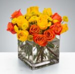 A vibrant floral arrangement featuring bright yellow and orange roses mixed with yellow button poms in a clear square glass vase.