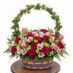 A vibrant floral arrangement in a colorful woven basket featuring red roses, pink tulips, white alstroemeria, and green pompon chrysanthemums, topped with an ivy-wrapped handle.