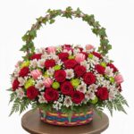 A vibrant and dense floral arrangement featuring red roses, pink tulips, and white alstroemeria in a colorful woven basket with a green ivy-covered handle, set against a white background.