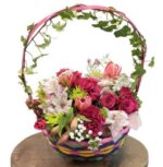 A vibrant floral arrangement in a colorful woven basket featuring pink roses, white alstroemeria, and a decorative ivy hoop handle, resting on a wooden surface.