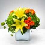A vibrant floral arrangement featuring bright yellow lilies, orange spray roses, and green hydrangea in a modern clear square glass cube vase.