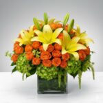 A lush and vibrant floral arrangement featuring bright yellow lilies, orange spray roses, and green hydrangeas in a square glass cube vase.