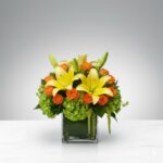 A vibrant floral arrangement featuring bright yellow lilies, orange spray roses, and green hydrangeas in a square glass cube vase.
