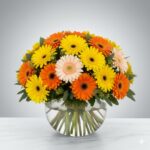 A vibrant bouquet of yellow, orange, and pale pink Gerbera daisies arranged in a clear glass fishbowl vase on a white marble surface.