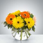 A glass fishbowl vase filled with vibrant orange and yellow gerbera daisies and fresh green foliage on a white marble surface.