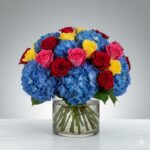 A vibrant and dense floral arrangement featuring blue hydrangeas, red roses, pink roses, and yellow roses in a clear glass cylinder vase filled with water, set against a neutral gray background.