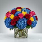 A vibrant and dense floral arrangement in a clear cylindrical glass vase, featuring large clusters of blue hydrangeas interspersed with red, yellow, and pink roses against a neutral grey gradient background.