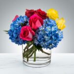A vibrant floral arrangement in a clear cylindrical glass vase featuring blue hydrangeas, pink roses, red roses, and yellow roses set against a clean white and gray background.
