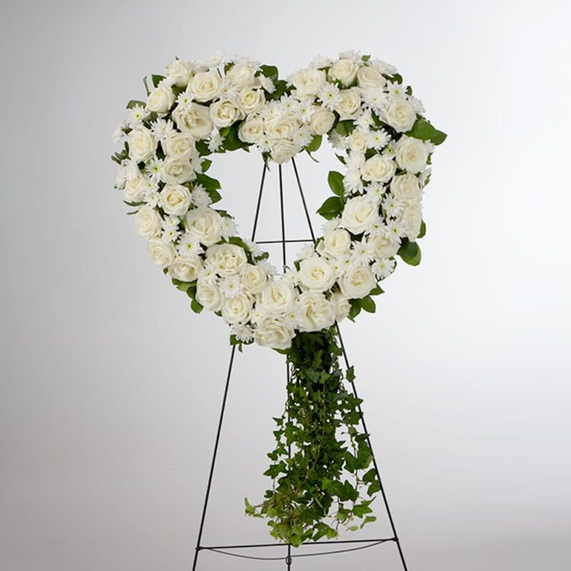 Heart-shaped white floral wreath with greenery on stand.
