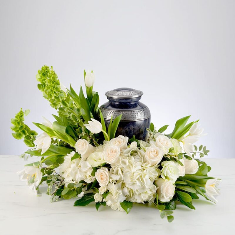A black urn surrounded by white flowers and green foliage on a white surface.