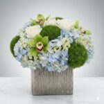 A modern floral arrangement in a square metallic silver vase featuring light blue hydrangeas, white roses, lime green cymbidium orchids, and spherical green trick dianthus (moss-like flowers).