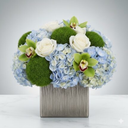 A sophisticated floral arrangement in a metallic silver square vase, featuring light blue hydrangeas, white roses, green cymbidium orchids, and decorative green moss balls.