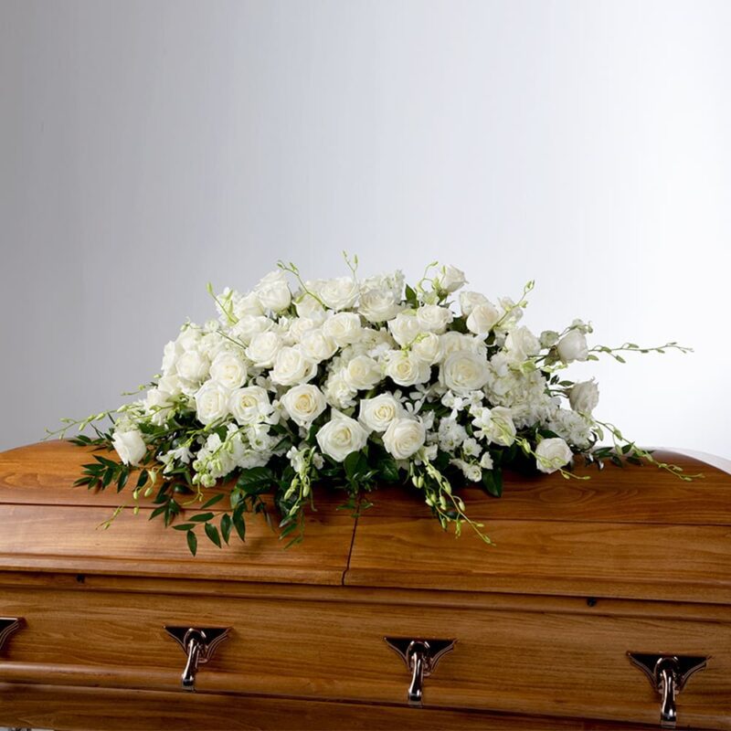 A large, elegant casket spray of white roses and lilies arranged on a polished wooden casket.