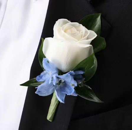 A close-up of a elegant wedding boutonniere featuring a single white rose and small blue delphinium accents pinned to a black suit lapel.