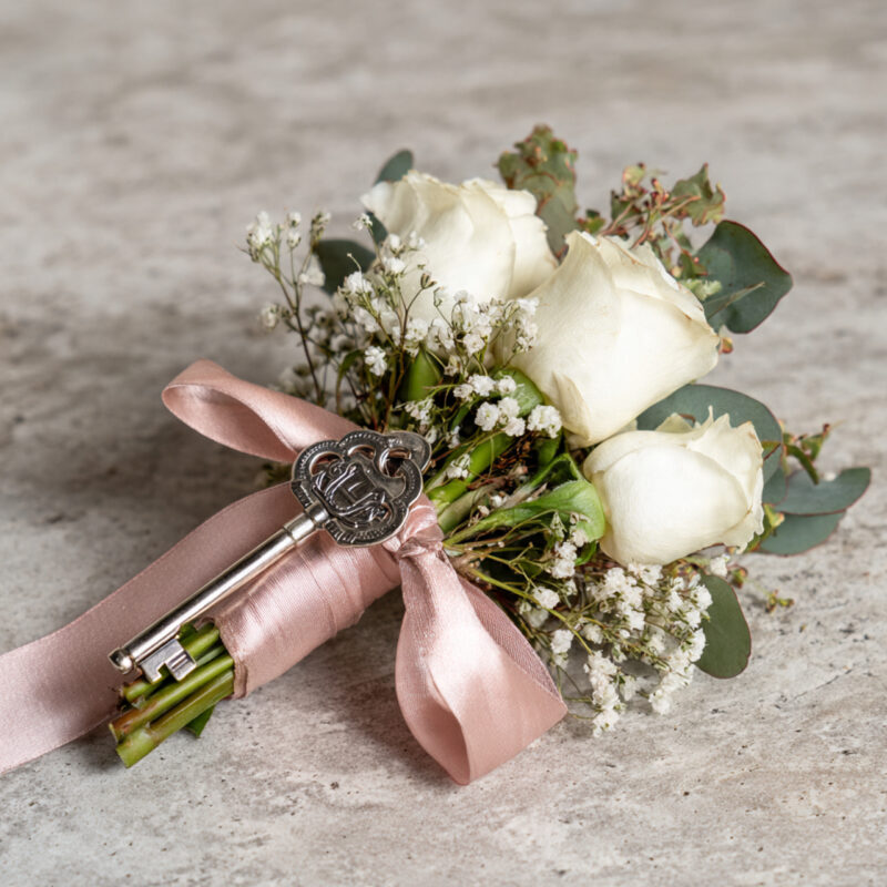 Small bouquet of three white roses with baby’s breath and eucalyptus, wrapped in blush pink satin ribbon and accented with an ornate silver key charm, on a light stone surface.