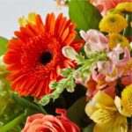 A vibrant autumn flower bouquet featuring a large orange Gerbera daisy, pink snapdragons, yellow alstroemeria, and small yellow button chrysanthemums with lush green foliage.