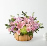 A lush floral arrangement in a light wood woven basket featuring pink lilies, pink roses, carnations, purple stock flowers, and a vibrant green spider mum, set against a clean white background.