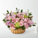 A lush floral arrangement in a light wood woven basket featuring pink lilies, pink roses, light purple stock flowers, and green spider mums accented with dark green foliage.