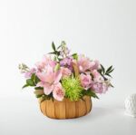 A round woven wooden basket filled with a soft pastel floral arrangement featuring pink lilies, pink roses, light purple stock flowers, and a vibrant lime green spider mum, set against a clean white background.