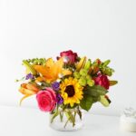 A vibrant and colorful mixed flower bouquet in a clear glass cylinder vase, featuring a large sunflower, bright pink roses, orange lilies, and purple accent flowers against a plain white background.