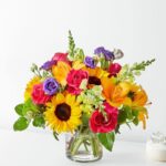 A vibrant and colorful floral bouquet in a clear glass vase, featuring sunflowers, pink roses, orange lilies, and purple lisianthus.