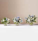 Three glass vases of varying sizes containing "Clear Skies" flower bouquets with blue hydrangeas, white roses, and silvery-green foliage, arranged in a row against a neutral beige background.