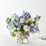 A lush floral arrangement in a clear glass cylinder vase featuring light blue delphiniums, white roses, light blue hydrangeas, white pompon mums, and dusty miller greenery set against a minimalist white background.