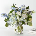 A premium floral arrangement in a glass vase featuring white roses, light blue hydrangeas, blue delphiniums, white pompon mums, and dusty miller foliage.